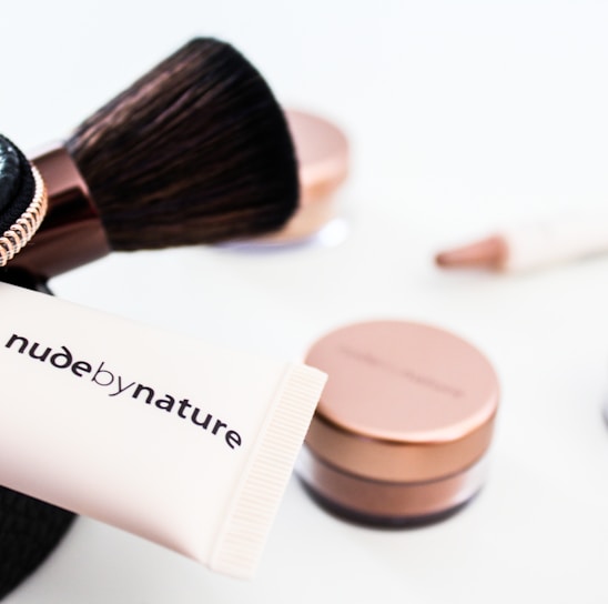 A collection of cosmetics is neatly displayed, including a makeup brush, a black zippered pouch, and several rose-gold capped jars. A tube that reads 'nude by nature' is partially visible, suggesting a beauty product brand.