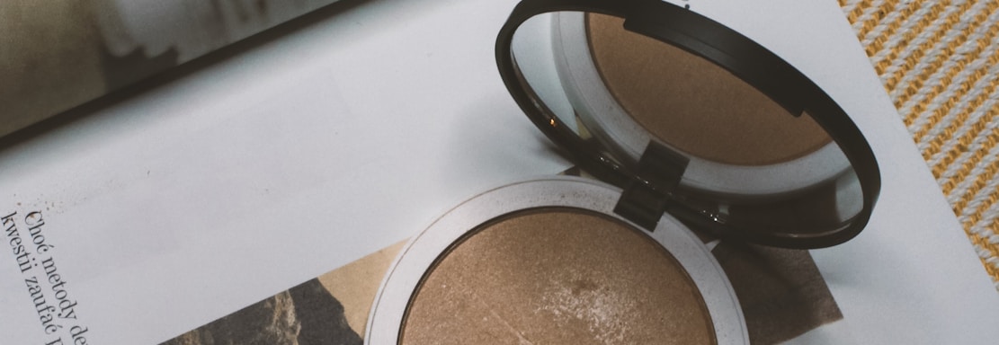 A makeup compact and brush rest on the pages of an open magazine. The magazine features an image of a woman with wavy hair looking back over her shoulder at the camera. Text is visible on the pages, and the overall setting creates a sense of everyday beauty and style.